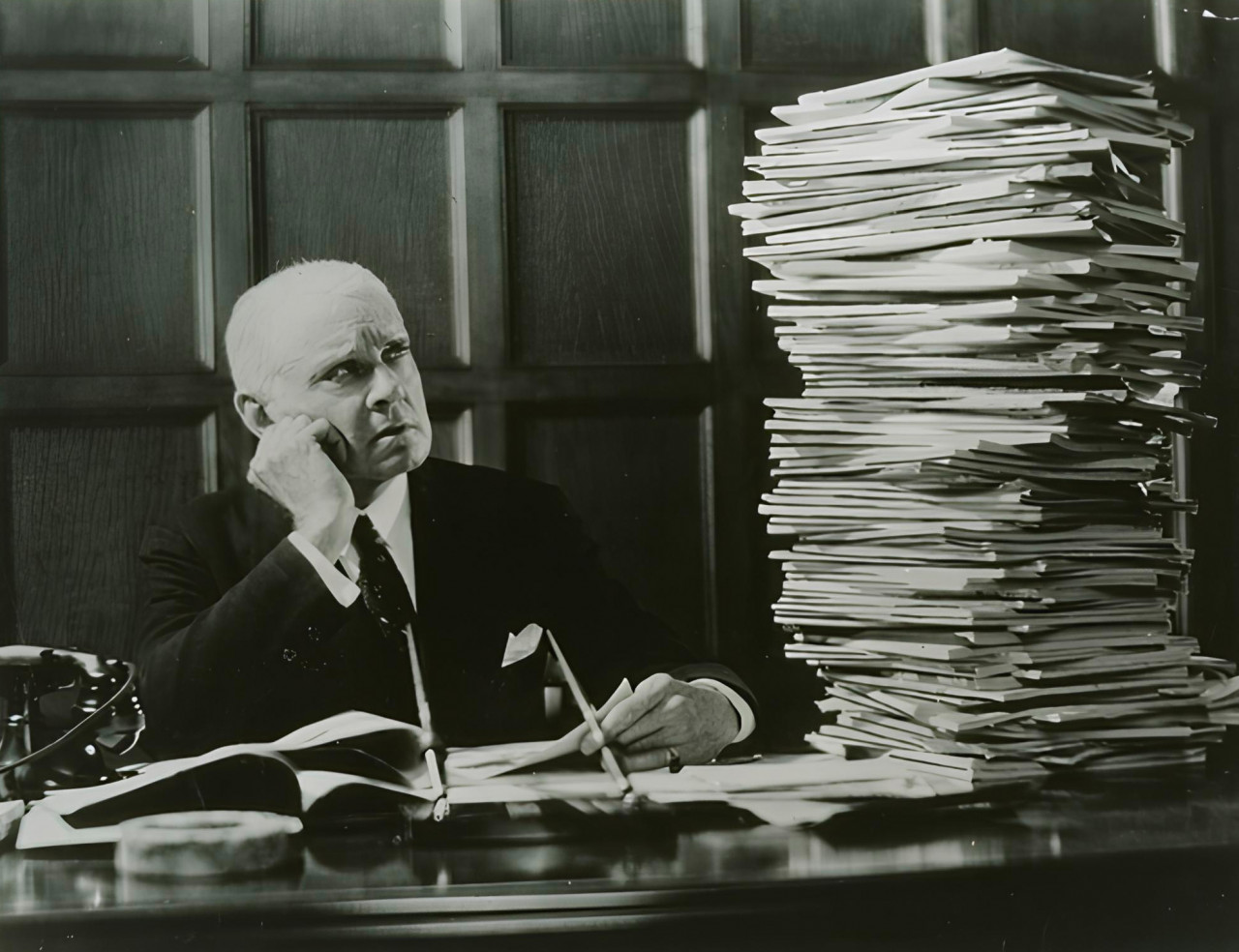 An old black and white photograph of a man at a desk, staring at a huge stack of papers.
