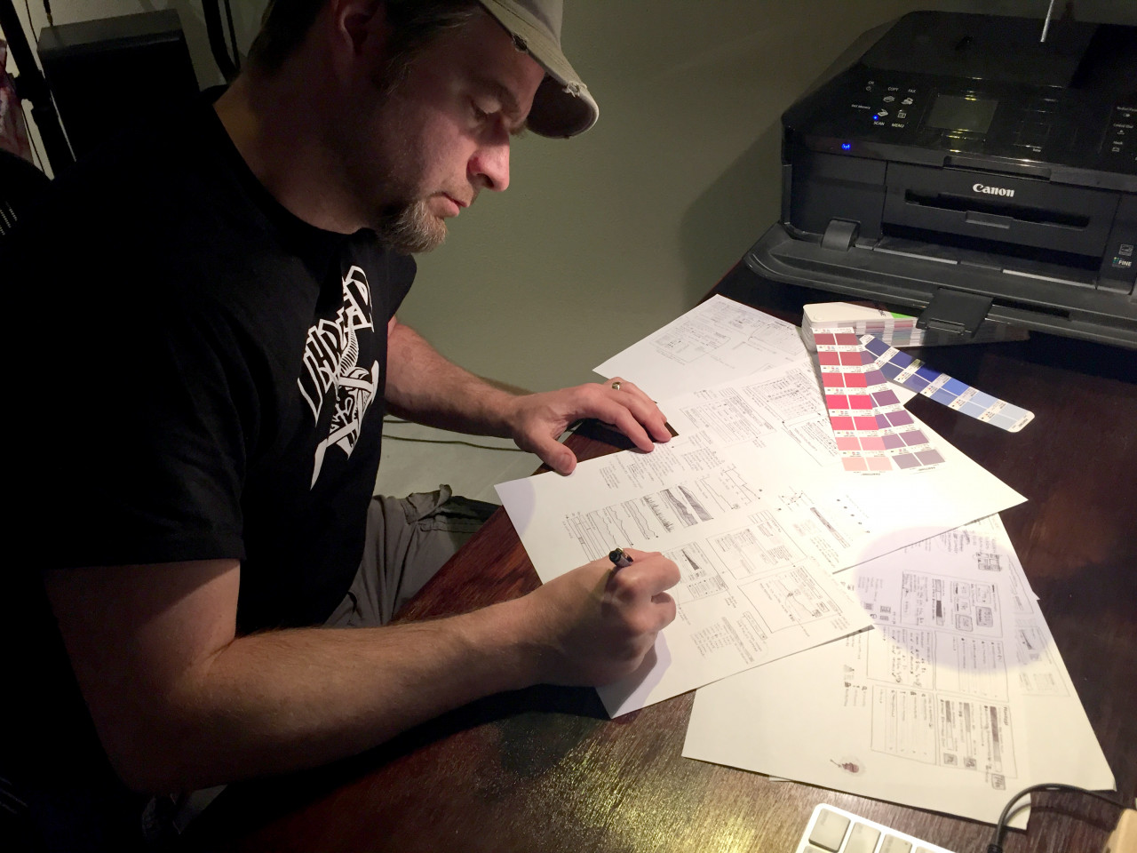 A picture of Chris at a desk, sketching with pen and paper. A fan deck of color swatches has several colors open in the background.